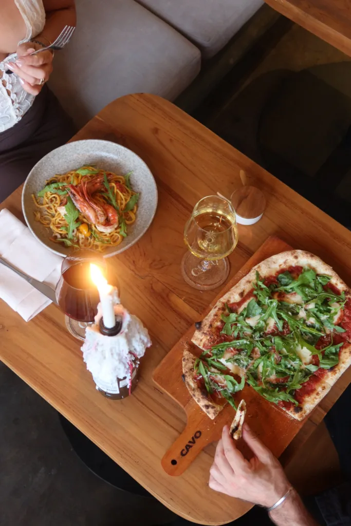 Overhead view of an oval Roman-style pizza with rocket, prawn spaghetti, wine and a candle on a table for two at CAVO Uluwatu